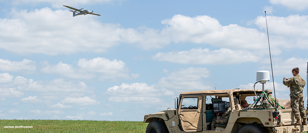 Army soldiers sitting in a Humvee in an open field as a large military plan flies overhead.