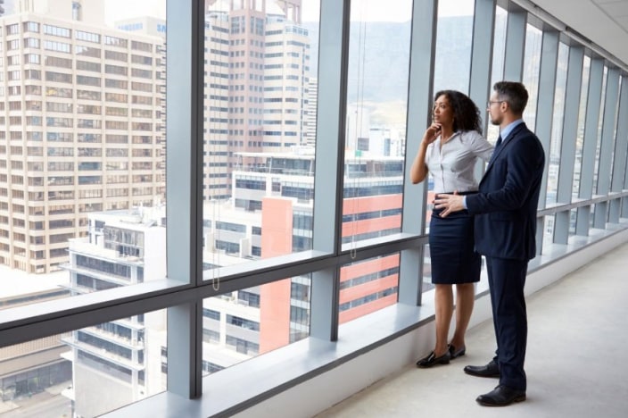A male and female coworker standing and looking out a large window in an office building.